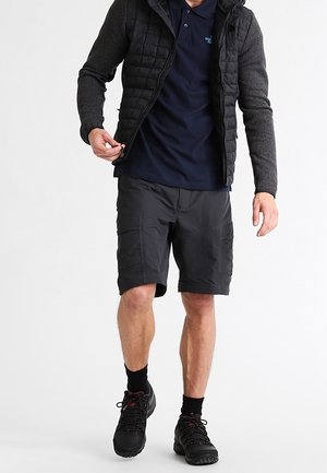Hombre con chaqueta acolchada negra, camiseta polo azul marino, pantalones cortos de carga oscuros, calcetines negros y zapatos de senderismo negros, ajustando la cremallera de la chaqueta.