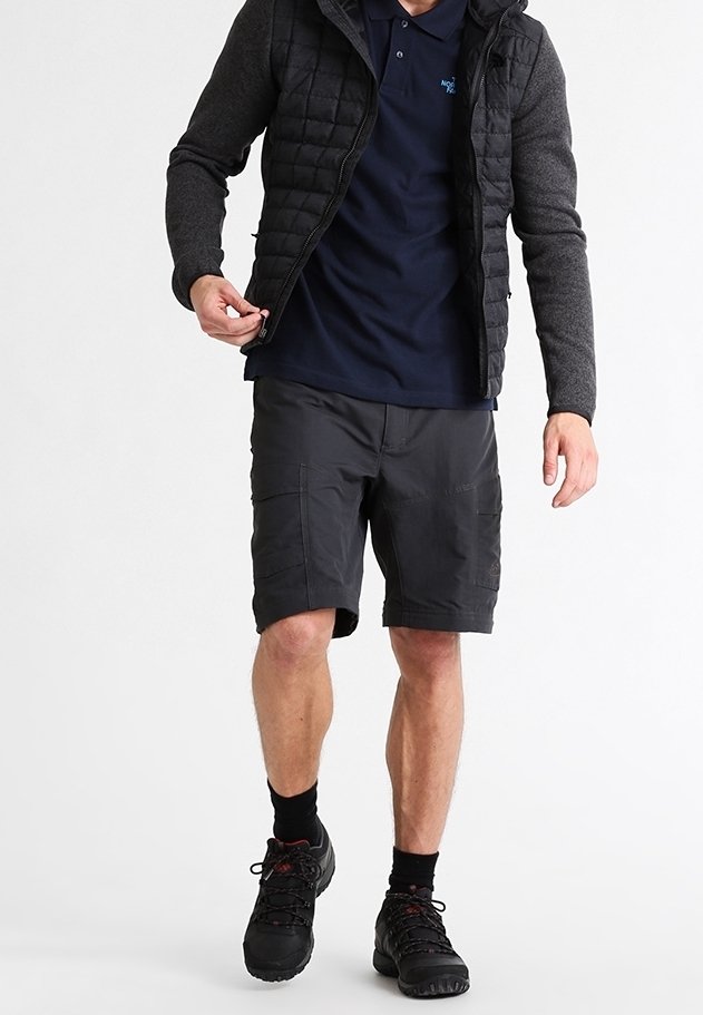 Mies, joka on pukeutunut mustaan tikattuun takkiin, tummansiniseen pikeepaitaan, tummiin cargo-shortseihin, mustiin sukkiin ja mustiin vaelluskengiin, säätää takin vetoketjua.