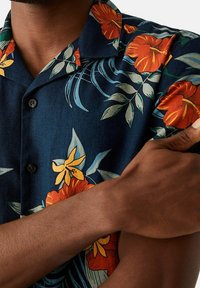 Camicia a maniche corte con bottoni in tessuto blu scuro, caratterizzata da una stampa floreale con fiori arancioni e gialli, foglie verdi e una consistenza liscia.