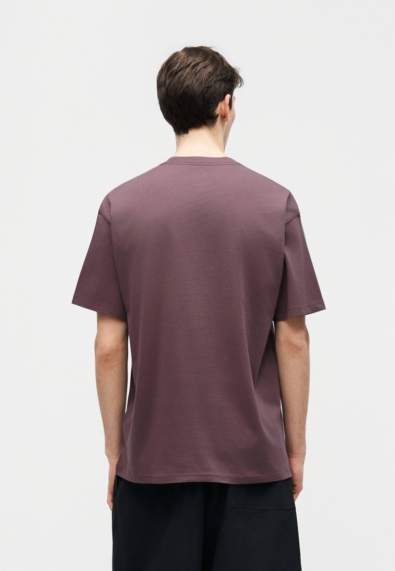 Ein Mann steht mit dem Rücken zur Kamera, trägt ein einfarbiges, kurzärmeliges T-Shirt in Mauve und schwarze Hosen vor einem neutralen Hintergrund.