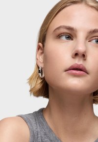 Los pendientes de aro de plata presentan un pequeño charm negro. La modelo tiene el cabello corto y rubio, y lleva una camiseta sin mangas gris.