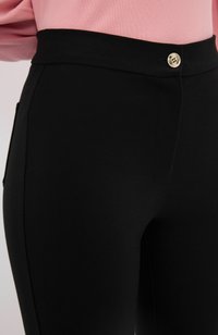 Zwarte hoge taillebroek met een gladde textuur, voorzien van een gouden knoopsluiting en zijzakken voor functionaliteit.