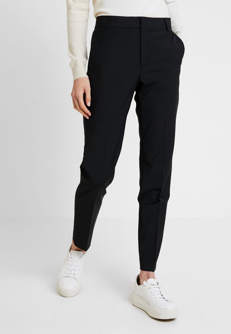 Schwarze maßgeschneiderte Hose aus glattem Stoff, mit schmalem Schnitt, flachem Bund und seitlichen Taschen. Kombiniert mit weißen Sneakers.