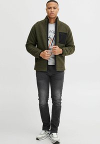 Olivegrøn fleecejakke med sorte detaljer, ståkrave og lynlåslomme. Båret over en grå graphic t-shirt og parret med mørke jeans.