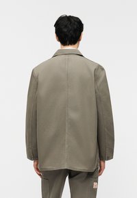 Άνδρας που φοράει ελιάς πράσινο μακρυμάνικο μπουφάν και ταιριαστά cargo παντελόνια, στέκεται με την πλάτη προς το φακό σε απλό λευκό φόντο.