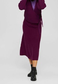 Personne portant une jupe portefeuille violette et un pull assorti avec des bottines noires, debout devant un fond gris clair uni.