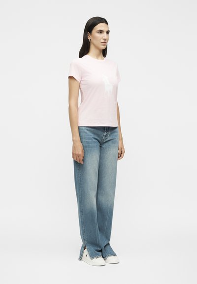 Γυναίκα με μακριά σκούρα μαλλιά, που φοράει ένα ανοιχτό ροζ κοντομάνικο μπλουζάκι, μπλε τζιν παντελόνι με φαρδιά μπατζάκια και σκισίματα, και λευκά αθλητικά, που στέκεται πλευρικά.