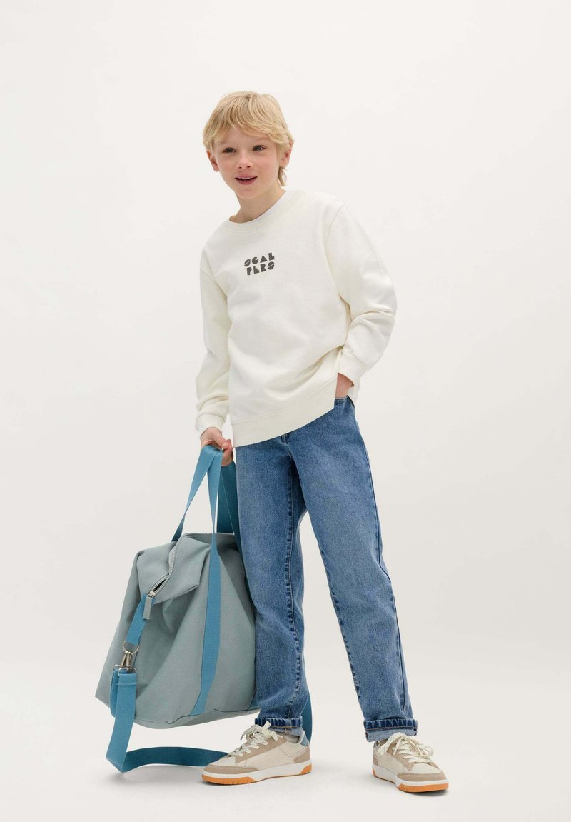 Felpa beige chiaro con scritte nere, jeans in denim blu e sneakers beige. Porta uno zaino di tessuto grigio con spallacci blu e una cerniera.