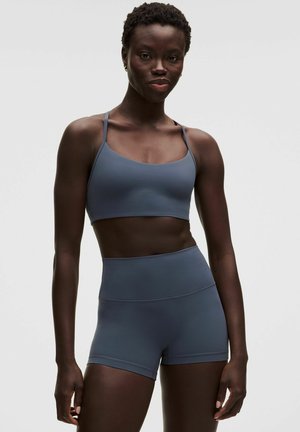 Atletisk kvinde iført en matchende blå sports-bh og shorts med høj talje, stående mod en ensfarvet lys baggrund.