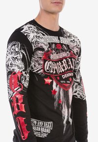 Schwarzes Langarmshirt mit grafischen Drucken in Weiß und Rot, einschließlich Totenköpfen, Text und Sternmotiven; aus dehnbarem Stoff gefertigt.