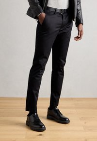 Pantalon noir en tissu lisse, présentant une coupe slim, des passants de ceinture et des poches latérales, associé à des chaussures noires à lacets.