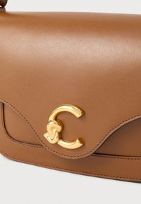 Borsa a mano in pelle marrone con design curvo e chiusura a ciondolo con la lettera "C" in tono oro. Texture liscia con dettagli di cuciture visibili.