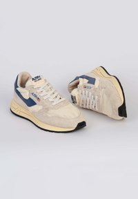 Sneakers beige con un mix di materiali in suede e tessuto, caratterizzate da un accento blu scuro e lacci bianchi. Suola texturizzata con dettagli neri.