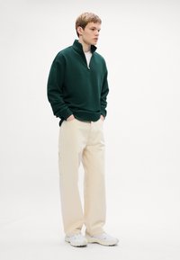 Sötét zöld, cipzáros pulóver magas gallérral, krémszínű cargo nadrággal és fehér sneakers-szel párosítva, egy modellen, sík háttér előtt.