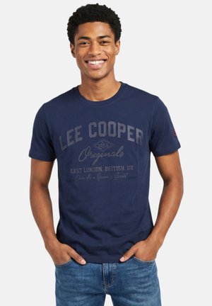Jeune homme souriant les mains dans les poches, portant un T-shirt Lee Cooper Originals bleu marine et un jean bleu, debout sur fond blanc.