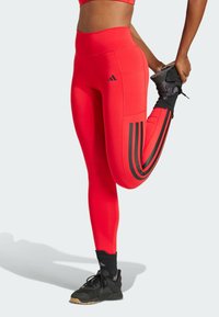 Leggings de sport rouges en matière extensible, avec un design à trois rayures noires sur le côté et une taille haute. Portés avec des chaussures de sport noires.