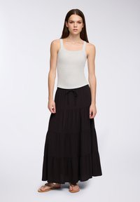 Jasnoszara, dopasowana koszulka na ramiączkach o kwadratowym dekolcie, w połączeniu z czarną, warstwową spódnicą maxi. Modelka stoi boso na jasnym tle.