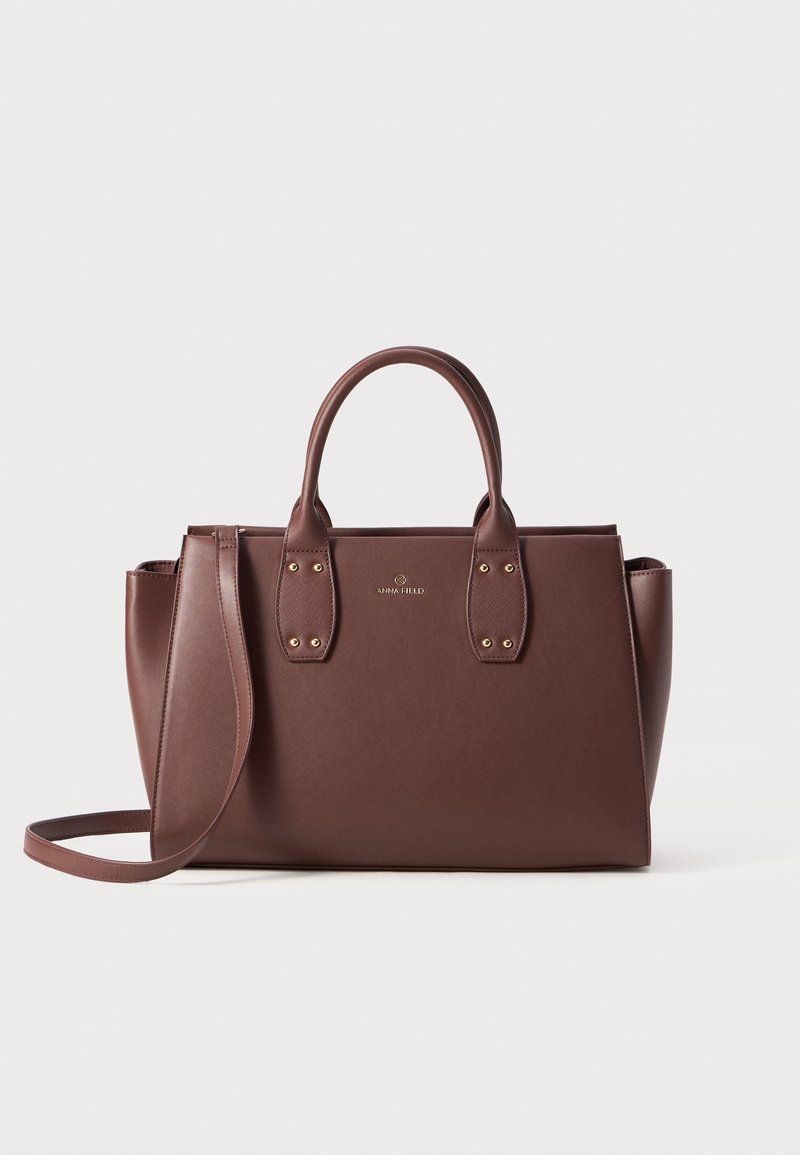 Sac à main en cuir marron avec double poignée, bandoulière amovible et accessoires dorés subtils sur fond uni.