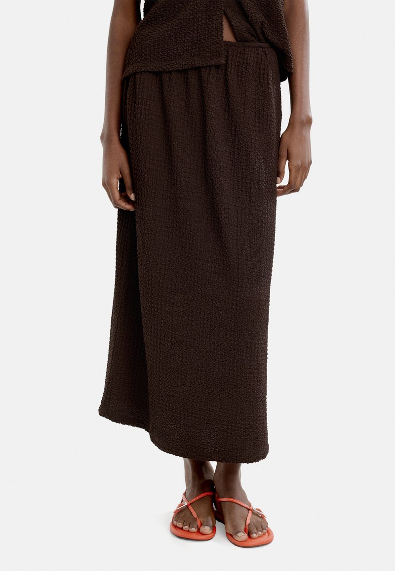 Brąskowy, teksturowany spódnicą o luźnym kroju, z nałożonym wzorem i bocznym rozcięciem, w zestawieniu z pomarańczowymi płaskimi sandałami z cienkimi paskami.