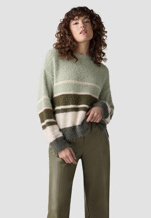 Junge Frau mit lockigem Haar trägt einen grünen, weißen und dunkel gestreiften flauschigen Pullover und olivgrüne Lederhosen vor grauem Hintergrund.