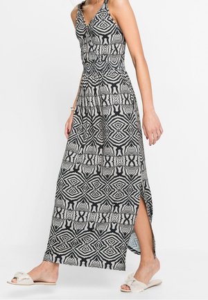 Donna che indossa un abito lungo senza maniche con motivo geometrico bianco e nero, con spacco laterale e sandali bianchi senza lacci.