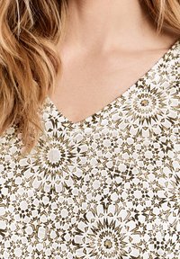 Top de cuello en V que presenta una tela blanca decorada con intrincados patrones geométricos en dorado y beige, exhibiendo un diseño texturizado.