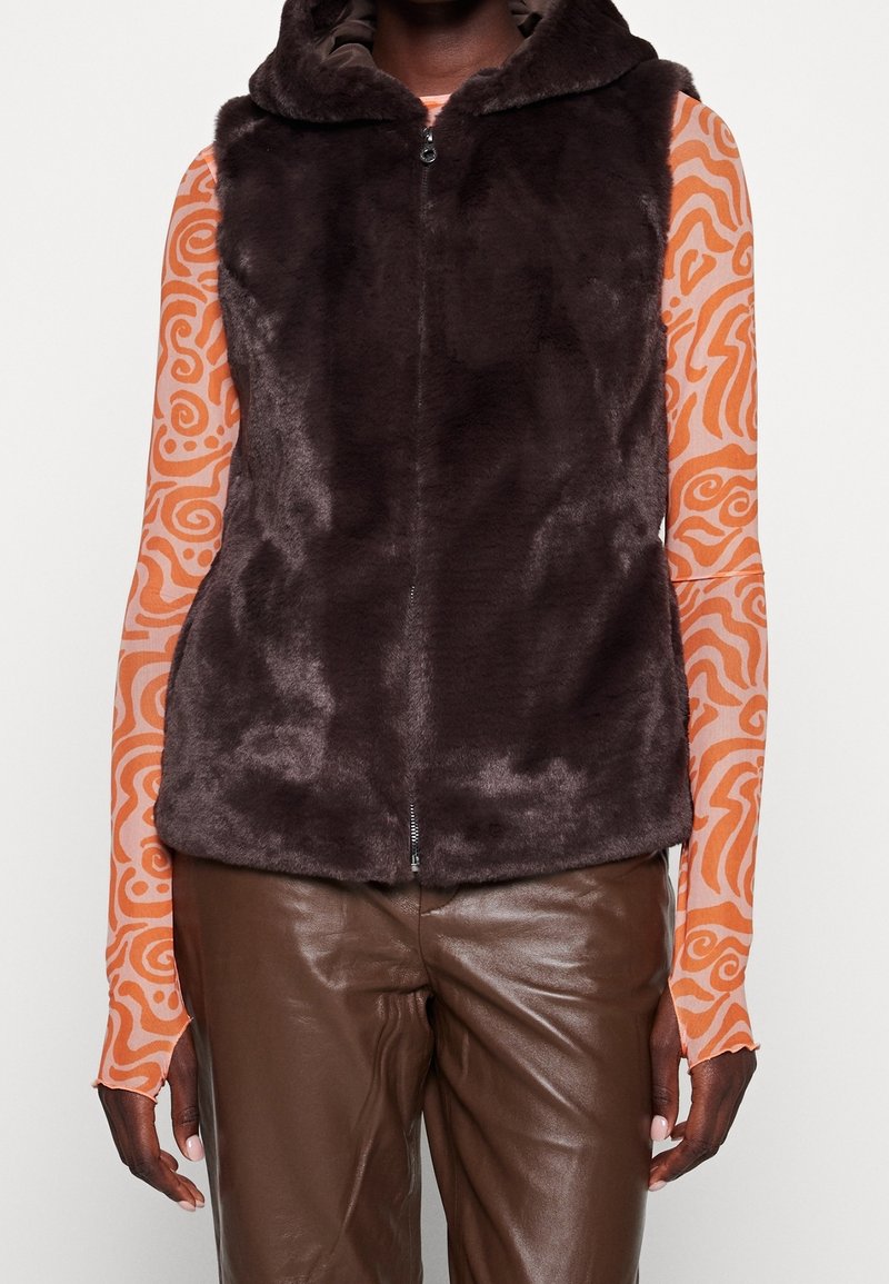 Dunkelbrauner Kunstfell-Weste mit Kapuze, Reißverschluss vorne und weicher Textur, getragen über einem gemusterten Langarmshirt und braunen Lederhosen.