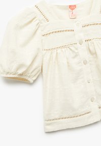 Blusa de algodón crema con mangas abullonadas, tejido texturizado, cierre frontal con botones y detalles de calado en el yugo y el dobladillo.