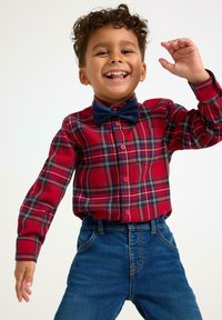 Smilende dreng i en rød plaid-skjorte med et marineblåt sløjfebånd og blå jeans, som løfter en hånd i en glad pose mod en hvid baggrund.