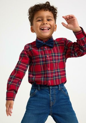 Uśmiechnięty chłopiec w czerwonej koszuli w kratę i granatowym muszniku, ubrany w niebieskie dżinsy, podnoszący jedną rękę w radosnej pozie na białym tle.