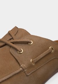 Κοντινή λήψη καφέ μοκασίνι από σουέντ με δερμάτινα κορδόνια περασμένα μέσα από χρυσά κρικάκια και λεπτομερή ραφές.