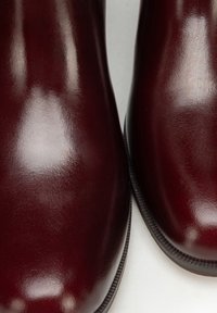 Bordowe skórzane botki z połyskującym wykończeniem, zaokrąglonym noskiem i gładką fakturą. Ozdobione przeszyciami wzdłuż krawędzi podeszwy.
