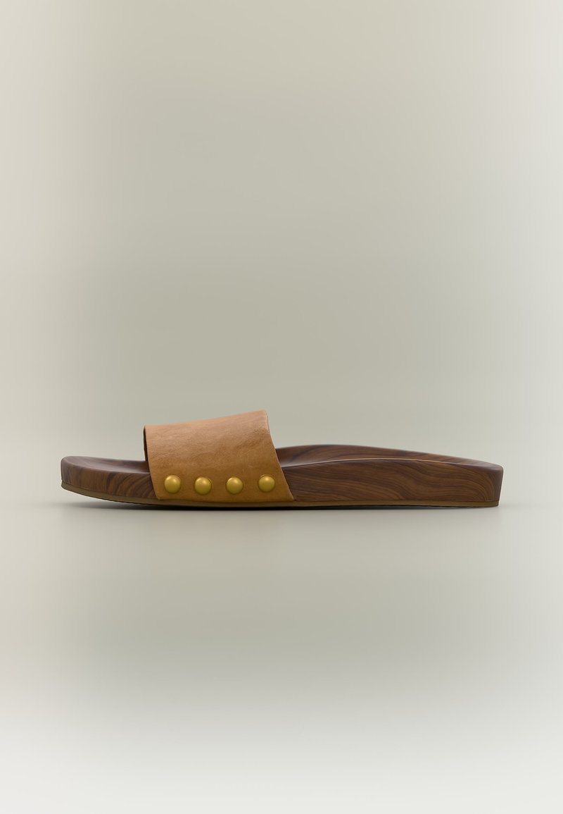 Sandale simple marron à enfiler avec une large lanière en cuir et quatre clous ronds dorés, semelle à texture boisée, fond minimaliste.