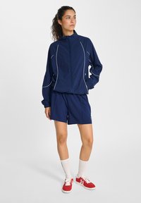Marinträningsdräkt med vit piping; zip-jacka och shorts; matchad med röda sneakers och vita sockor; atletisk design, lättviktsmaterial.