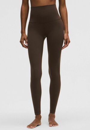 Egy nő magasderekú, sötétbarna, testhezálló leggingset visel, mezítláb áll egy egyszínű világos háttér előtt.