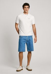 White polo shirt with a small repeating pattern, paired with blue denim shorts and white sneakers with dark accents. Casual outfit for warm weather.
