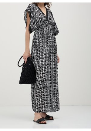 Femme portant une robe maxi sans manches à motifs noirs et blancs avec un col en V, tenant un sac à main tissé noir et portant des sandales plates noires.