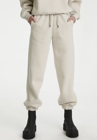Pantaloni della tuta beige in morbido materiale con polsini e vita elasticizzati. Presentano tasche laterali e un piccolo logo ricamato sul davanti.