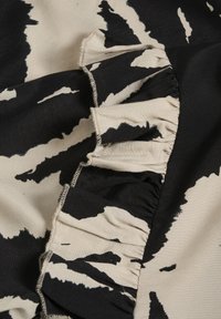 Tessuto nero e beige con un motivo audace, caratterizzato da volant lungo un bordo. La texture appare liscia e tessuta con cuciture visibili.