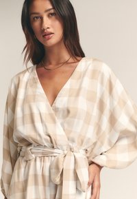 Smėlio spalvos ir baltos spalvos languota suknelė, rišama, su plačiomis rankovėmis, V formos iškirpte ir surišamu juosmens diržu, pagaminta iš glotnios, lengvos medžiagos.