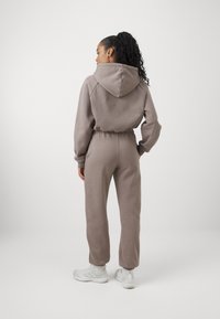 Bruine hoodie jumpsuit met elastische taille en taps toelopende enkels, gemaakt van zachte stof. Draagt met witte sportschoenen.