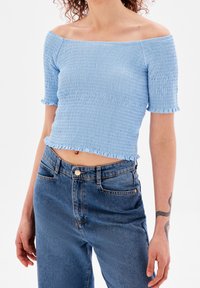 Světle modrý top bez ramínek se smockovanou texturou a krátkými, volánkovými rukávy. Kombinovaný s modrými džíny s vysokým pasem.