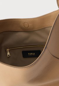 Interiorul geantii din piele camel cu un buzunar cu fermoar, căptușeală din material textil și o etichetă neagră pe care scrie „Furla din 1927.”