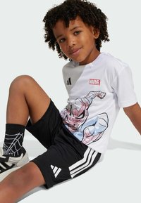 Bílá tričko s grafikou superhrdiny, v kombinaci s černými šortkami a ponožkami s motivem pavoučí sítě; ležérní sportovní oblečení pro děti.