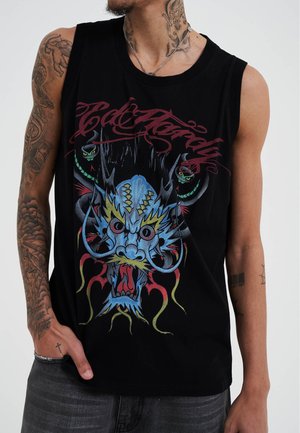 Un hombre tatuado viste una camiseta sin mangas negra con un gráfico colorido de un dragón, de pie contra un fondo liso.