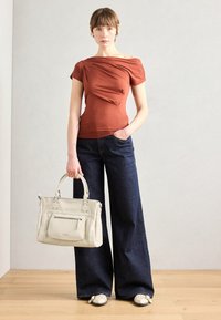 Haut à manches courtes plissé de couleur rouille, associé à un jean large bleu foncé. Le modèle tient un sac à main gris clair avec des accents métalliques. Sol en bois.