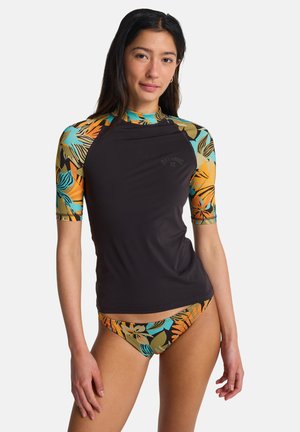 Frau trägt ein schwarzes Rashguard mit mehrfarbigen tropischen Bedruckten Ärmeln und passenden Bikinihosen, stehend vor einem weißen Hintergrund.