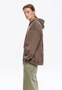 Sudadera marrón de gran tamaño con cordón, con una impresión gráfica en la parte posterior y bolsillo tipo canguro. Combinada con pantalones de pierna ancha de color verde claro.