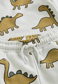 Hvide sweatsbukser har et gentaget dinosaurmotiv i brunt med grønne detaljer. Taljen har en elastisk og snorelukning.