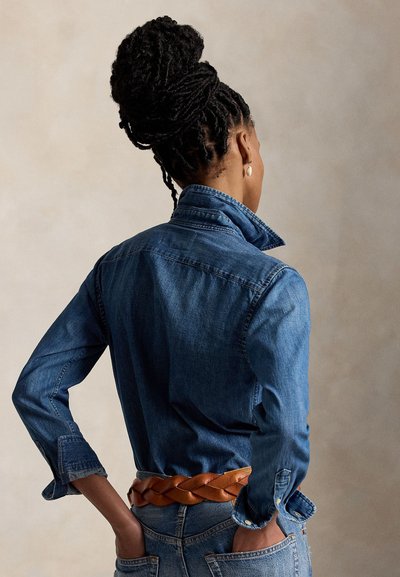 Vrouw met gevlochten haar in een knot, draagt een denimshirt in een spijkerbroek gestopt met een gevlochten bruine riem, staat met haar handen in de achterzakken.